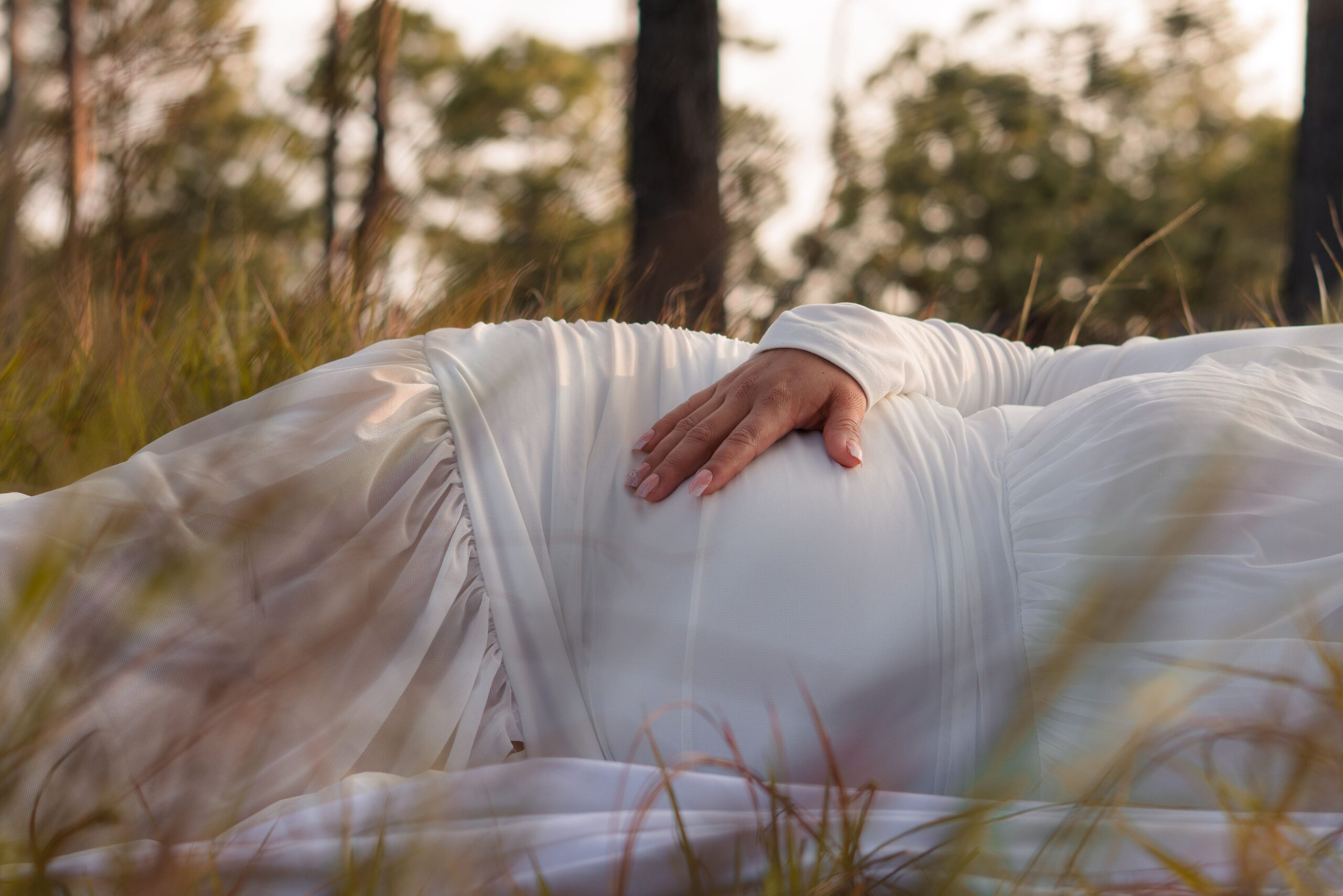 A peaceful outdoor maternity portrait with a pregnant woman in a white dress lying in the grass.
